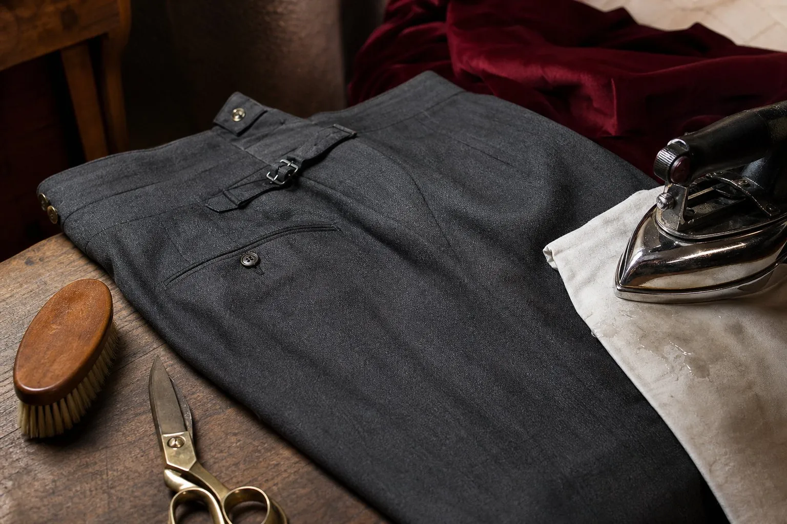 Pantalon de costume en laine grise posé sur une table de repassage d'atelier, avec pattemouille, fer et brosse à vêtements